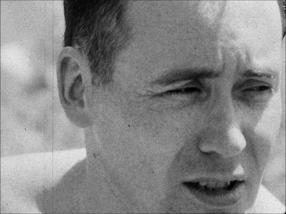 A film still of a close up of a man's face. He looks to the left, eyes half closed against the sun. His bare shoulders suggest he is shirtless.