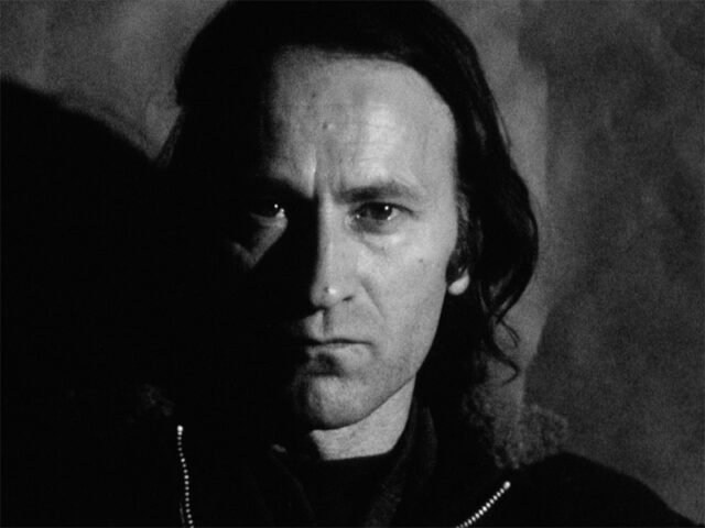 A film still of a close up of a man's head. He stares at the camera intensely with a hard expression, lips pushed together. The top of his jacket is visible, and the very top of his hair extends out of frame
