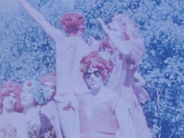 A film still. Multiple people stand together in the same wig and similar dresses. A person in center frame wears sunglasses, while three people to the left stand closely together. Three people stand on an elevation above the center person, dancing