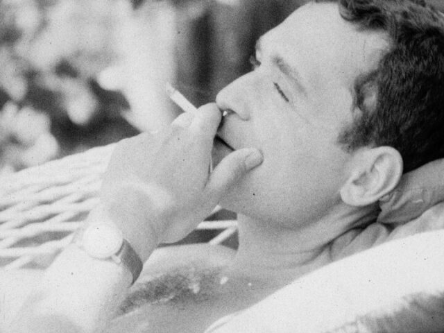 A black and white film still close-up a man smoking a cigarette while laying down in a hammock. His eyes are closed while his fingers hold the cigerette. Only the top of his hairy chest is viable.