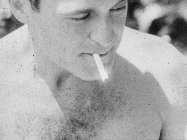 A black and white film still of a man smoking a cigarette. The top of his head is cropped out of frame, and the top of his hairy chest is visible.