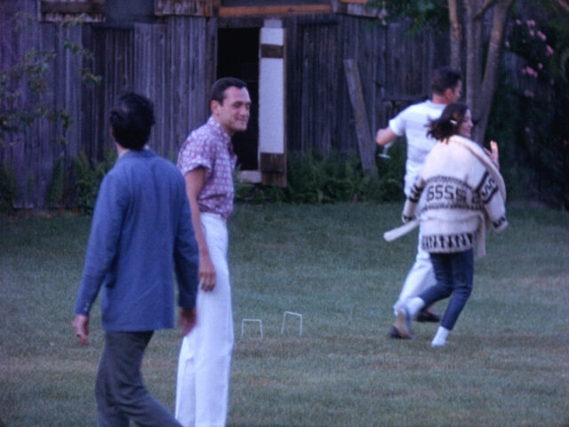 A film still of people in a yard. Two men stand on the left of the frame, while a man and a woman run in on the right. A wine glass is in the running man's hand.