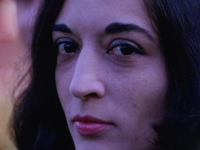 A film still of a woman looking at the camera. She had brown hair, red lipstick and slight blue eyeshadow. Her forehead and chin are slightly out of frame.