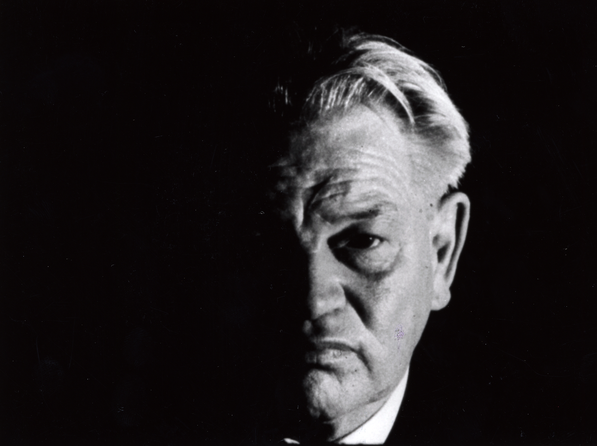 A black and white film still of a older-man's face. Half his face is obscured in shadow, while the other half has a serious expression. His hair is parted in the middle, pushing dramatically from the shadow