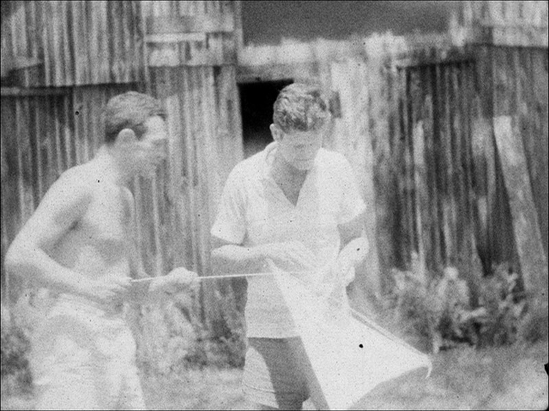 A grainy black and white photo of two men in front of a shed. The leftmost man is shirtless and is holding a string, while the other wears a white shirt and is holding a kite. Both look intently at the kite, seemingly repairing it.