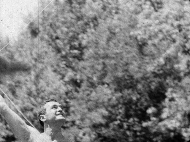 A film still with a man in the lower left corner flying a kite . He is shirtless, his right arm raised and looking at the kite off frame. The majority of the image is off trees in the back ground, out of focus.