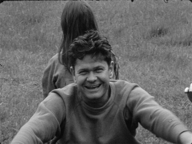 A black and white film still of a man sitting in grass looking up at the grass. He looks up at the camera, mid smile like he is laughing. His hands reach out to touch his knees, which are off camera. A person with long hair sits back-to-back with him, looking forward.