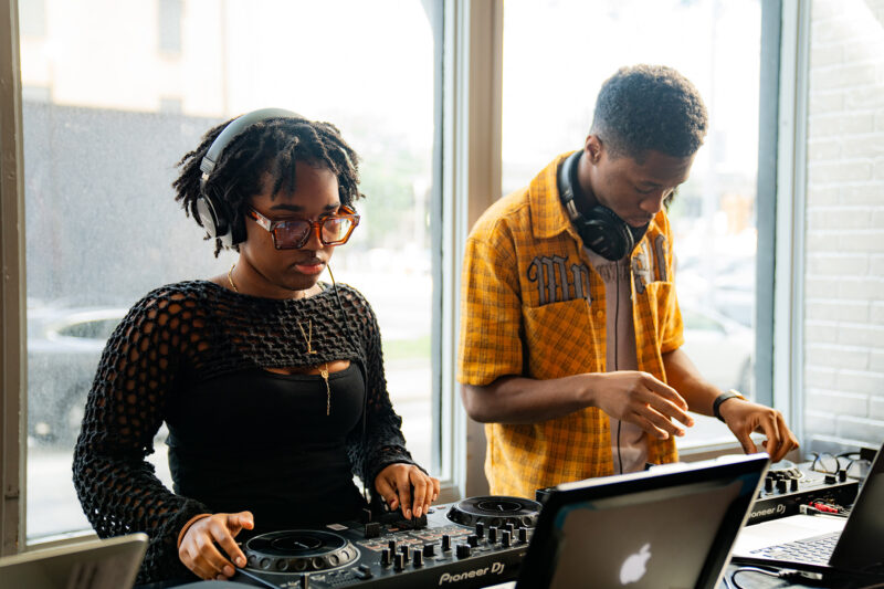 Two people using DJ equipment, with one person adjusting controls on a mixing deck and another selecting tracks on a laptop, in The Warhol entrance space.