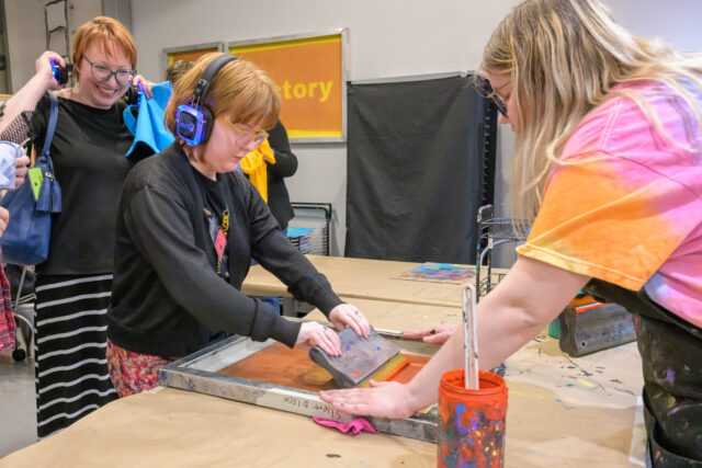Three individuals participate in a screen printing workshop at The Andy Warhol Museum, using a squeegee to apply ink on a screen frame. Various art supplies and vibrant colors are visible around them.