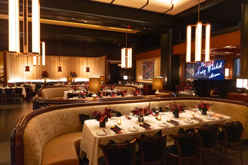 A photo of the interior of a fancy restaurant. The lighting is warm and inviting, and each table is long and lavishly set with flowers, candles, and a pamphlet. On the right, a screen reads Andy Warhol in cursive script.