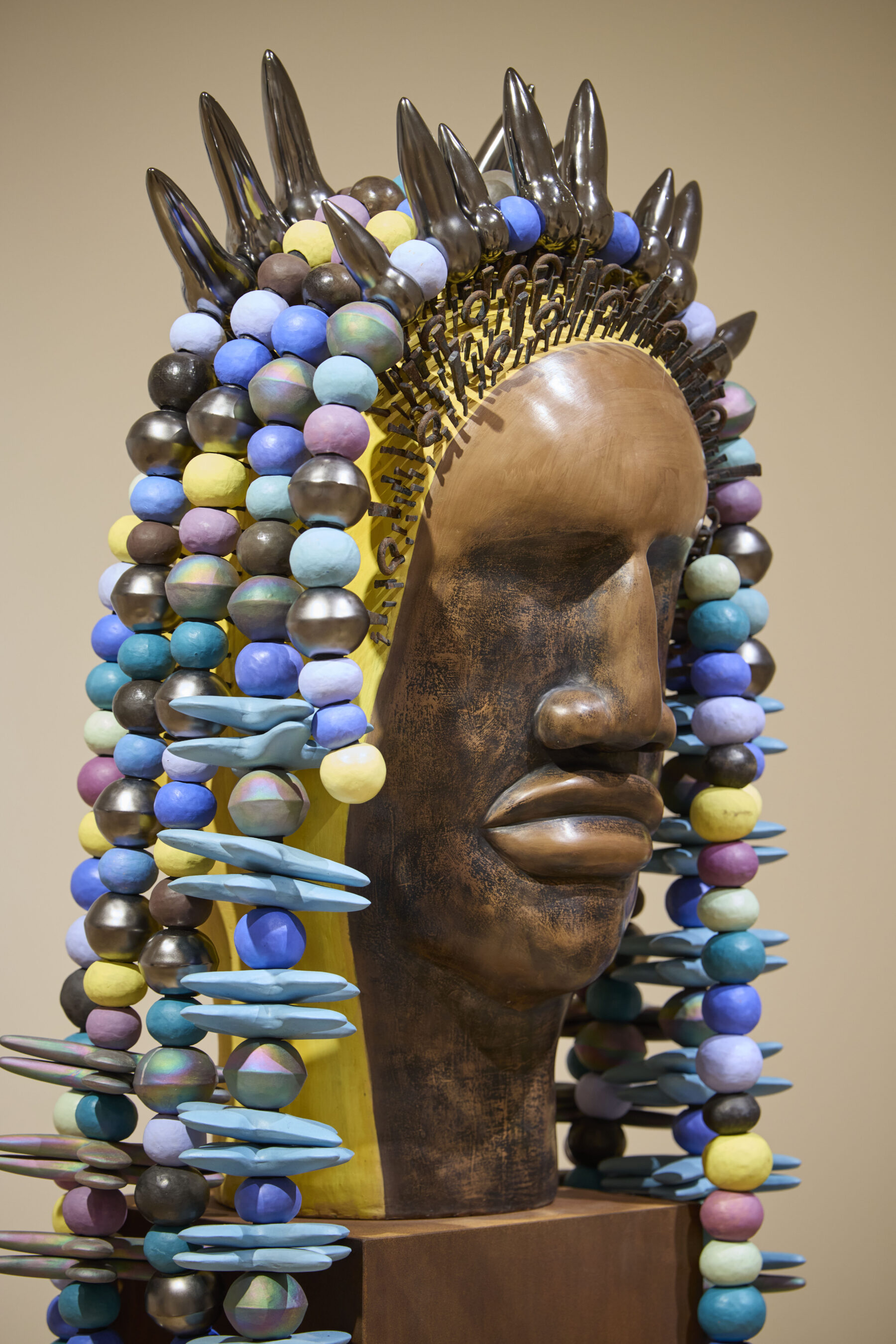 A colorful ceramic sculpture of a human head with a smooth, bronze-like surface. The head is adorned with a striking headdress composed of jagged spikes and vibrant beads in various colors, including blue, purple, and yellow. The sculpture stands on a metal base against a neutral background, highlighting its intricate details and textures