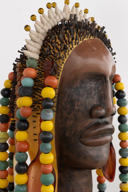 A detailed ceramic sculpture of a human head with a dark, polished surface. The head features an elaborate headdress made of intricate spikes and colorful beads, including orange, yellow, green, and black. Adorning the headdress are lighter-colored decorative elements. The sculpture is presented against a plain white background, emphasizing its textures and vibrant colors.