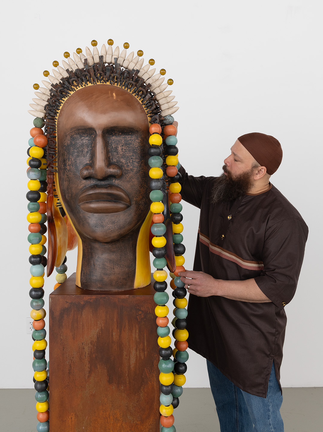 An artist with a beard, wearing a brown shirt and a tan cap, is adjusting a large, colorful ceramic sculpture of a human head. The sculpture features a polished dark surface and an elaborate headdress adorned with vibrant beads in various colors. The artist is positioned beside the sculpture, which stands on a rust-colored base against a plain white background, highlighting the details of both the sculpture and the artist.