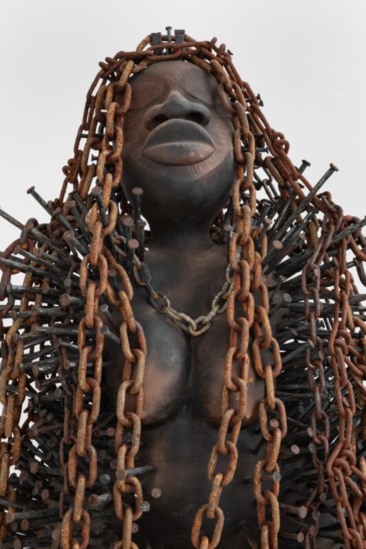 A detailed ceramic sculpture of a human chest and head with a dark, polished surface. The head features dreadlocks made of rusted chains, secured by old nails to the head. A mass of nails stab the statue around the body, the chains interlacing with some. The photo is taken from navel level and tilted upwards to see the sculpture's face