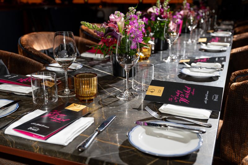 A photo taken looking down a long marble dinner table, just above a corner. The table is set for a fancy dinner. Each setting has a black pamphlet reading Andy Warhol in pink, cursive text. The table is decorated with floral arrangements and candles regularly spaced down the middle.