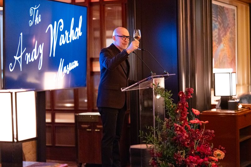 Mario R. Rossero, director of The Warhol, stands behind a glass podium toasting with a wine glass. He is wearing a black suit, and stands infront of a screen reading The Andy Warhol Museum in cursive script