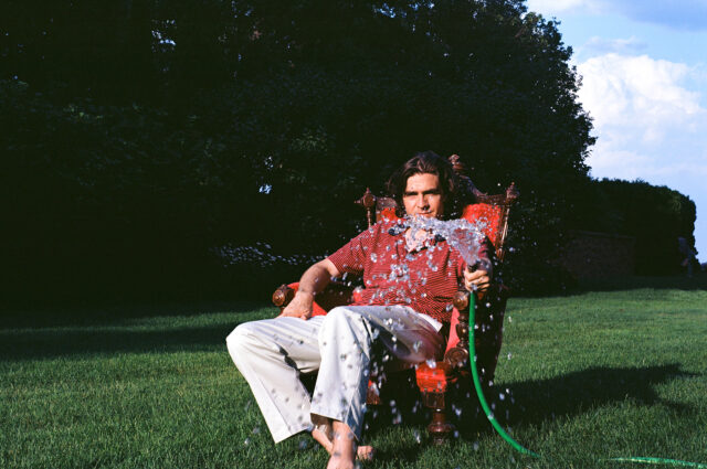 Person sitting on a red chair outdoors, spraying water from a garden hose on themself, with green grass and trees in the background.