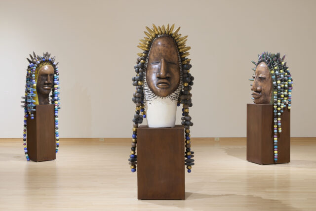 3 detailed sculptures of a human head with a dark, polished surface are arranged on top of pedestals in a gallery with a white wall. The center pedestal is closer to the camera and facing the user, while the other two are turned to face the center mask. Each sculpture features an elaborate headdress made of intricate spikes and colorful beads, adorned with light colored decorative elements.