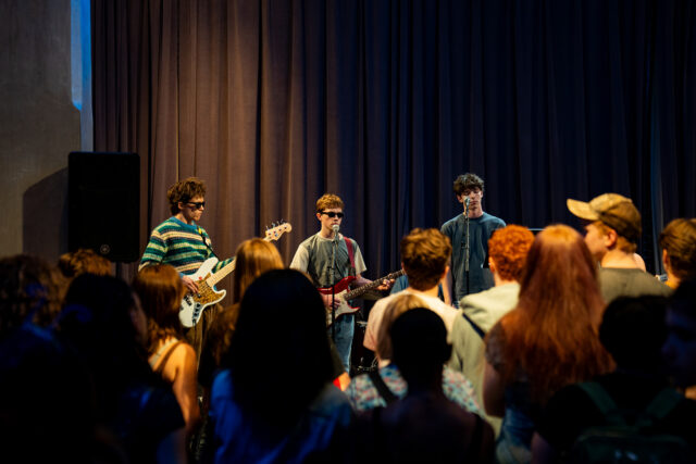 A music band is performing on stage in front of a crowd. The band members are playing guitars and standing in front of a dark curtain. The audience is facing the band.