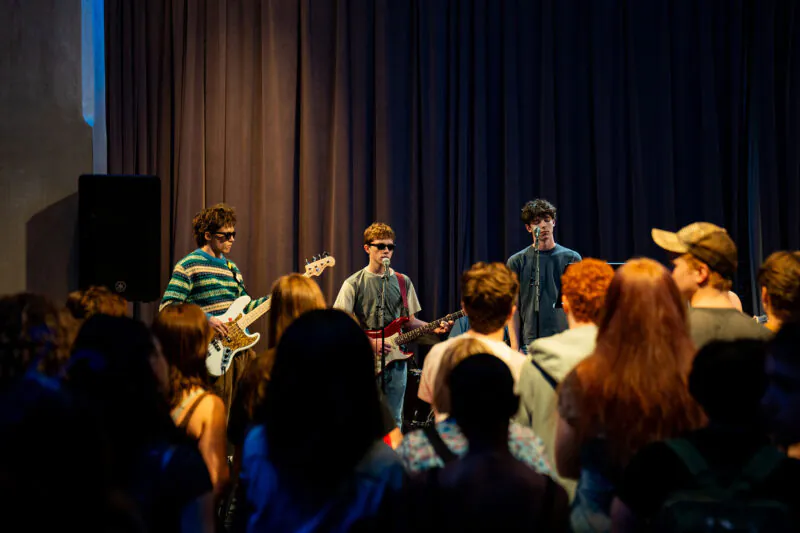 A music band is performing on stage in front of a crowd. The band members are playing guitars and standing in front of a dark curtain. The audience is facing the band.
