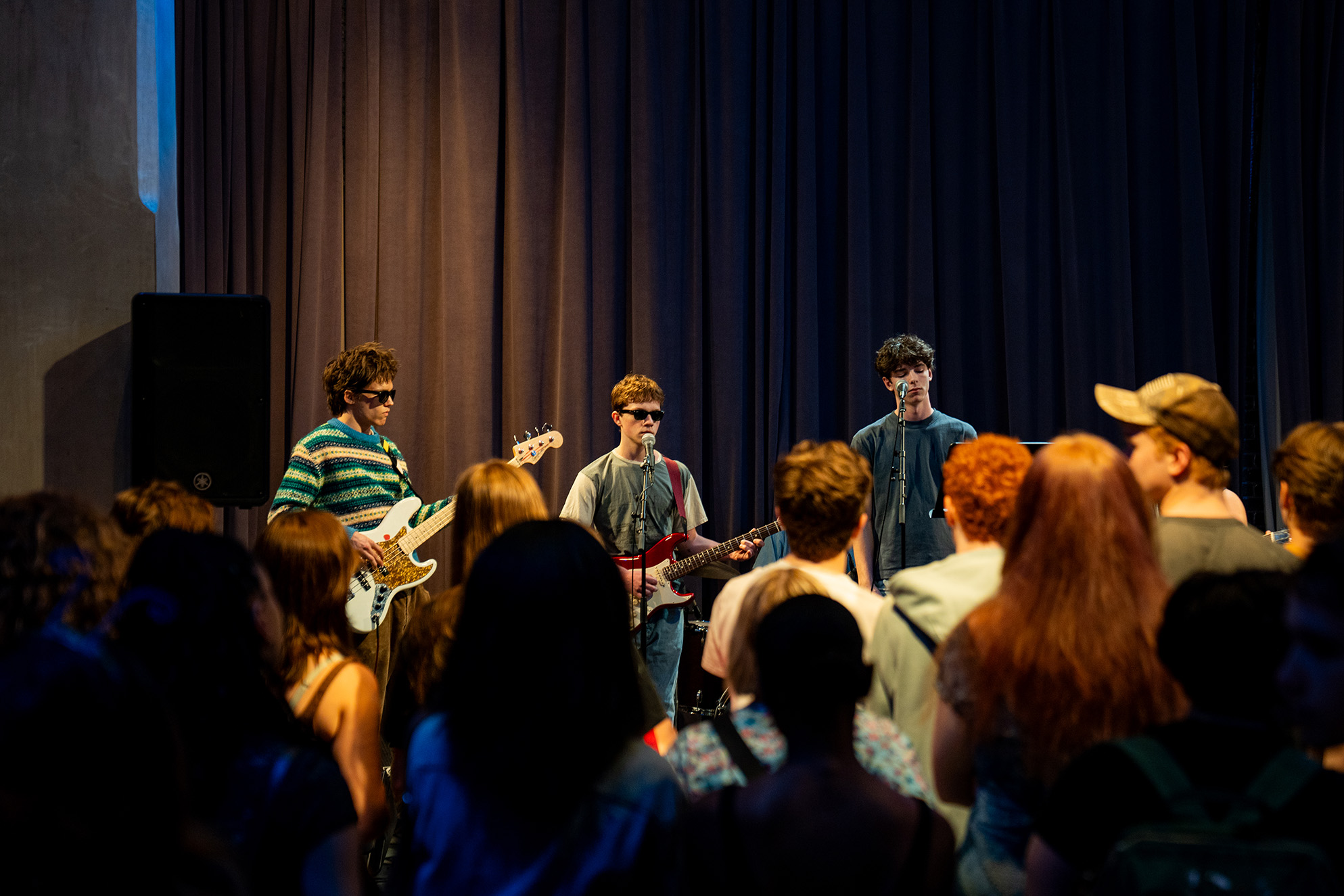 A music band is performing on stage in front of a crowd. The band members are playing guitars and standing in front of a dark curtain. The audience is facing the band.