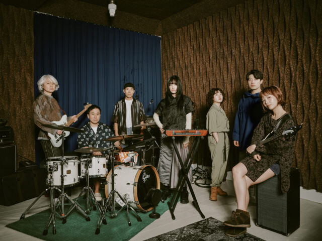 A band is posing in a music studio with various instruments, including drums, keyboards, and guitars. The room has textured walls and a blue curtain. Seven band members are standing and seated, with some holding musical instruments.