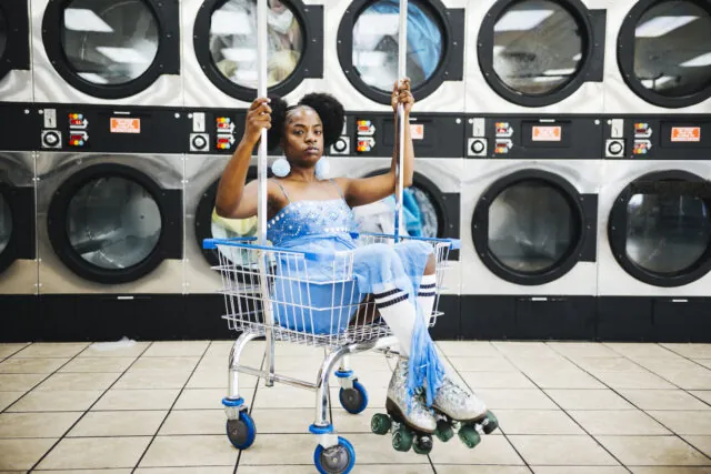 Sunny War in a shimmering blue outfit and roller skates sitting in a cart on wheels at a laundromat.