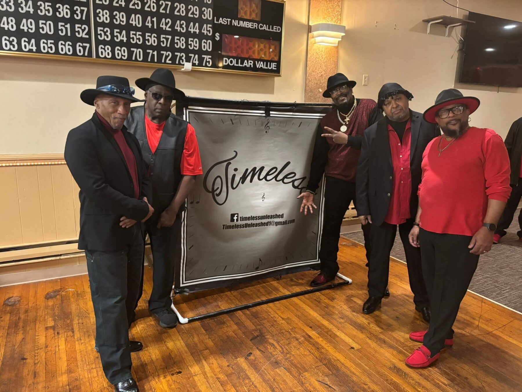 A group of individuals dressed in coordinated red and black attire stands in front of a Timeless banner. They are in a room with a wooden floor and a wall displaying a number board in the background.