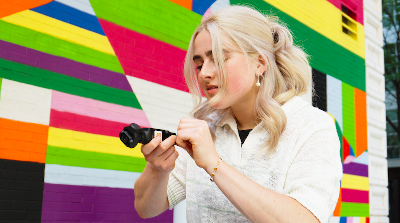 The image features an individual standing in front of a vibrant, multicolored striped wall. The person is holding a camera, pointing it towards the direction of the viewer. The person has blonde hair and is wearing a white top.