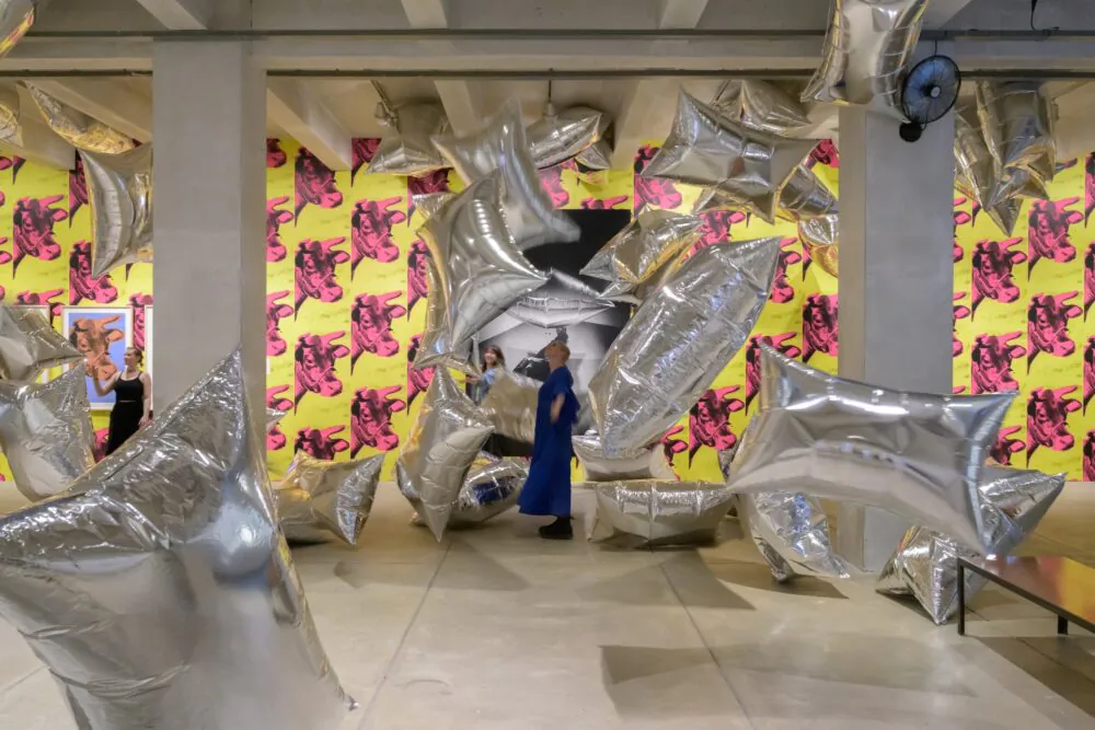 A room with cement ceiling and floor, the back wall is covered in wallpaper depicting pink cow heads on a yellow background. A woman stands in-front of a large picture, looking up at large pillow shaped balloons surrounding her.