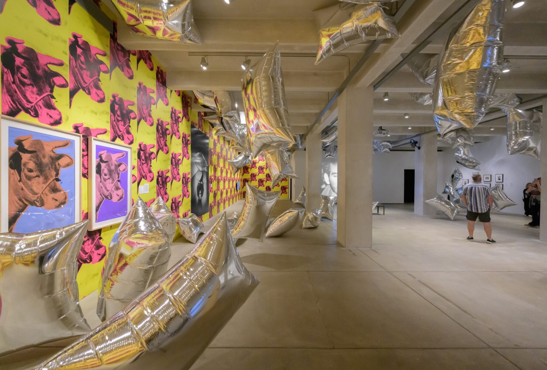 A art gallery with cement ceiling and floor, the left wall is covered in wallpaper depicting pink cow heads on a yellow background while the right wall is a clean white. Two people observe large reflective pillow shaped balloons drifting across the room.