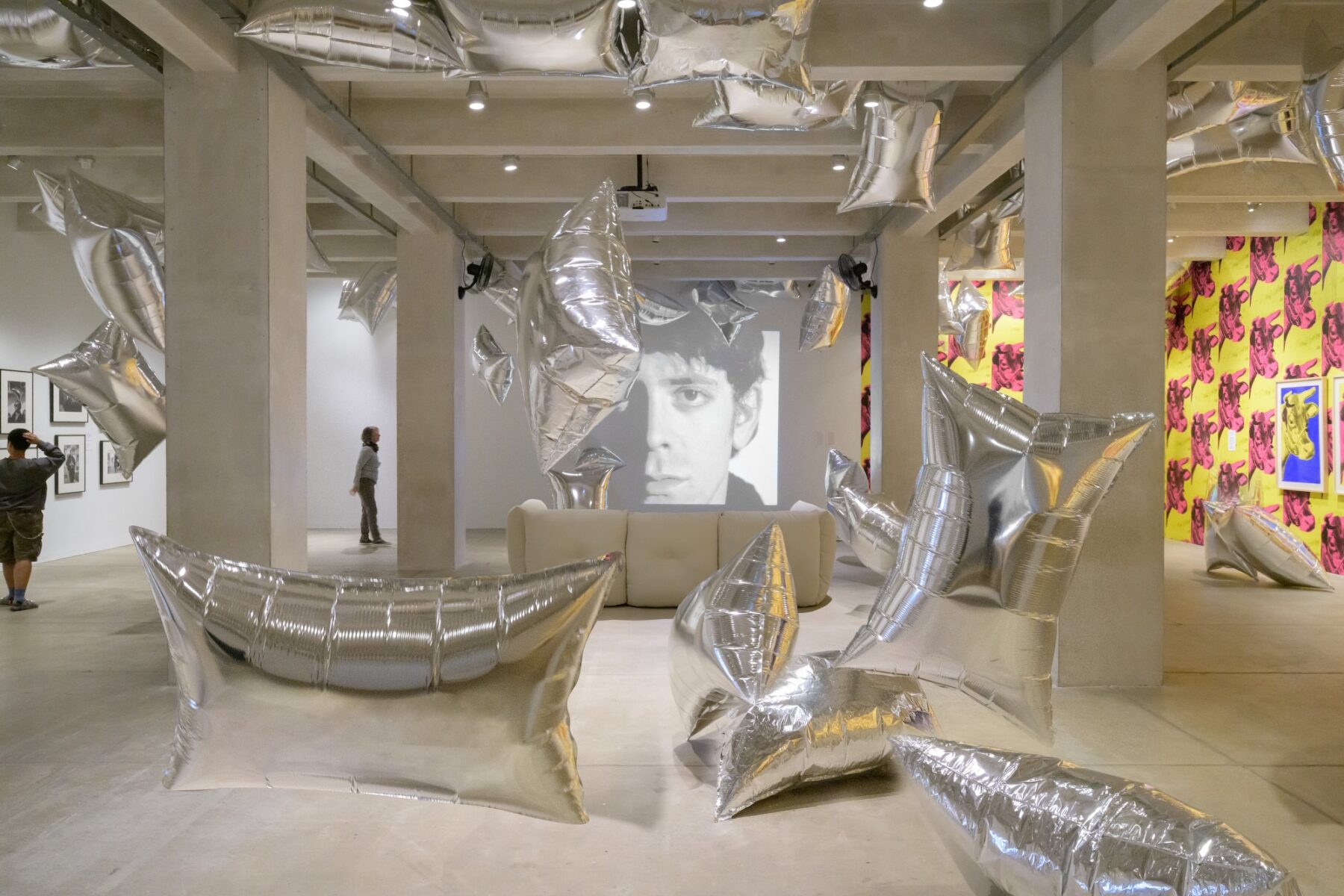 An art gallery with cement ceiling and floor with large silver pillow shaped balloons drifting around the room. A black and white video of a man's face staring at the camera is projected onto the far wall, and is framed by white structural support pillars and a white couch facing the projection.