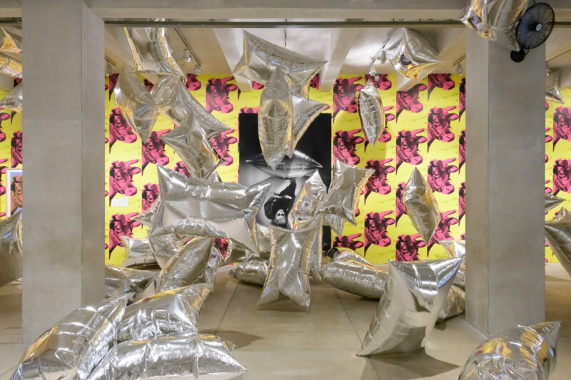 A room with cement ceiling and floor, the back wall is covered in wallpaper depicting pink cow heads on a yellow background. there is a black and white photo of Andy Warhol laying on the ground centered on the wall. In the foreground are several silver rectangular balloons floating about.