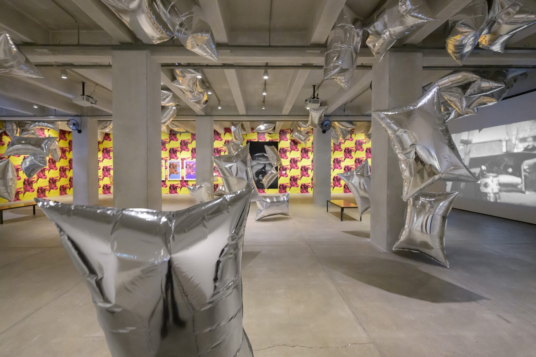 A wide shot of a room with cement ceiling and floor, the back wall is covered in wallpaper depicting pink cow heads on a yellow background. The room is full of large reflective pillow shaped balloons drifting freely, while the walls display a picture of Andy Warhol laying down and a projected black and white movie.