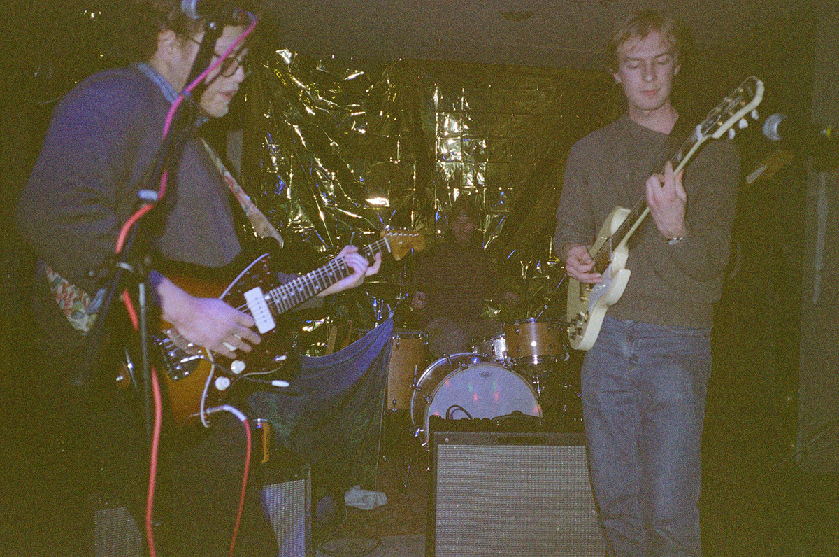 Three musicians performing on stage with guitars and a drum set. A metallic backdrop is visible behind them. The setting appears to be a small venue.
