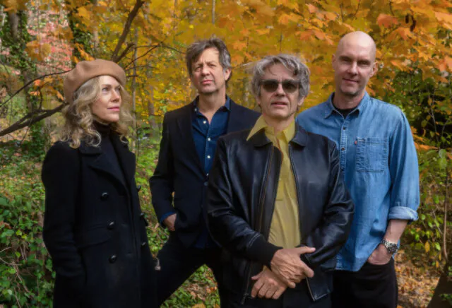 A group of four adults standing outdoors in an autumn setting with colorful foliage. They are dressed in casual to smart-casual attire, with one wearing a beret and another in a leather jacket. The scene is serene and natural, with vibrant fall colors in the background.