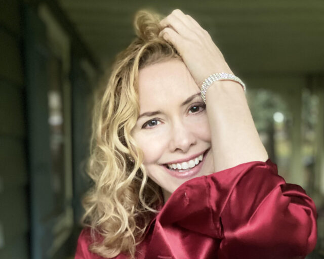 Portrait of Nellie McKay smiling wearing a red blouse. Her left hand is up on her forehead in her shoulder length blonde hair.