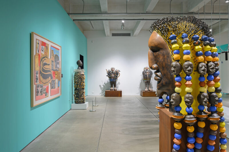 An art gallery at The Andy Warhol Museum featuring vibrant, eclectic sculptures and artwork. On the left, a wall displays a colorful mixed-media piece. In the center and right, several large sculptures with intricate designs made from various materials are exhibited. The sculptures include human-like figures adorned with colorful beads and textured surfaces, positioned under well-lit conditions. The gallery has a spacious, industrial feel with concrete floors and high ceilings.