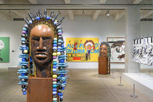 The image shows an art gallery at The Andy Warhol Museum with large wooden sculptures adorned with colorful beads. In the background, vibrant paintings are displayed on the walls. The artwork presents a mix of bold colors and abstract forms, contributing to a modern and eclectic atmosphere.