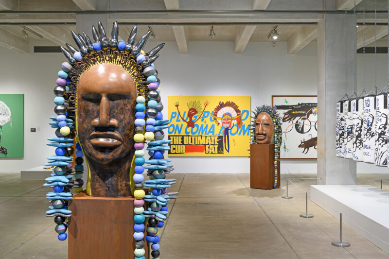 The image shows an art gallery at The Andy Warhol Museum with large wooden sculptures adorned with colorful beads. In the background, vibrant paintings are displayed on the walls. The artwork presents a mix of bold colors and abstract forms, contributing to a modern and eclectic atmosphere.