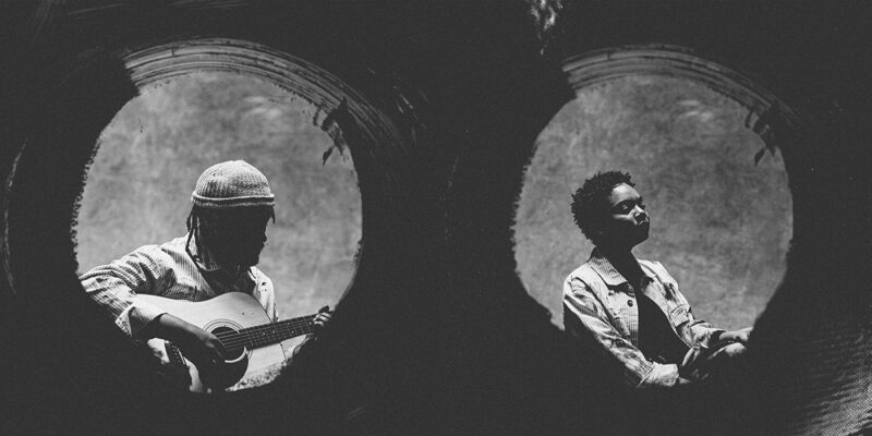 Two individuals are silhouetted inside circular frames. One is holding a guitar, and the other is wearing a jacket. The image is in black and white.