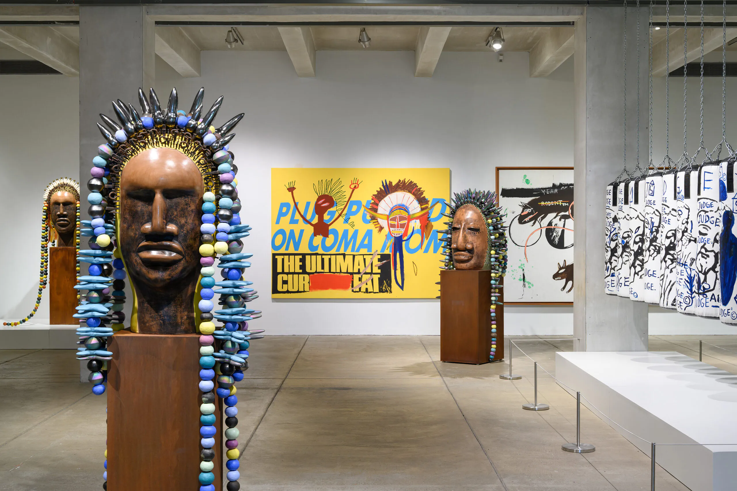 Contemporary art gallery featuring large sculptural heads on oxidized metal pedestals adorned with colorful bead-like elements, with bold graphic paintings on the walls by Jean Michel-Basquiat. A row of hanging white punching bags painted by Andy Warhol and Basquiat are suspended by chains on the right.