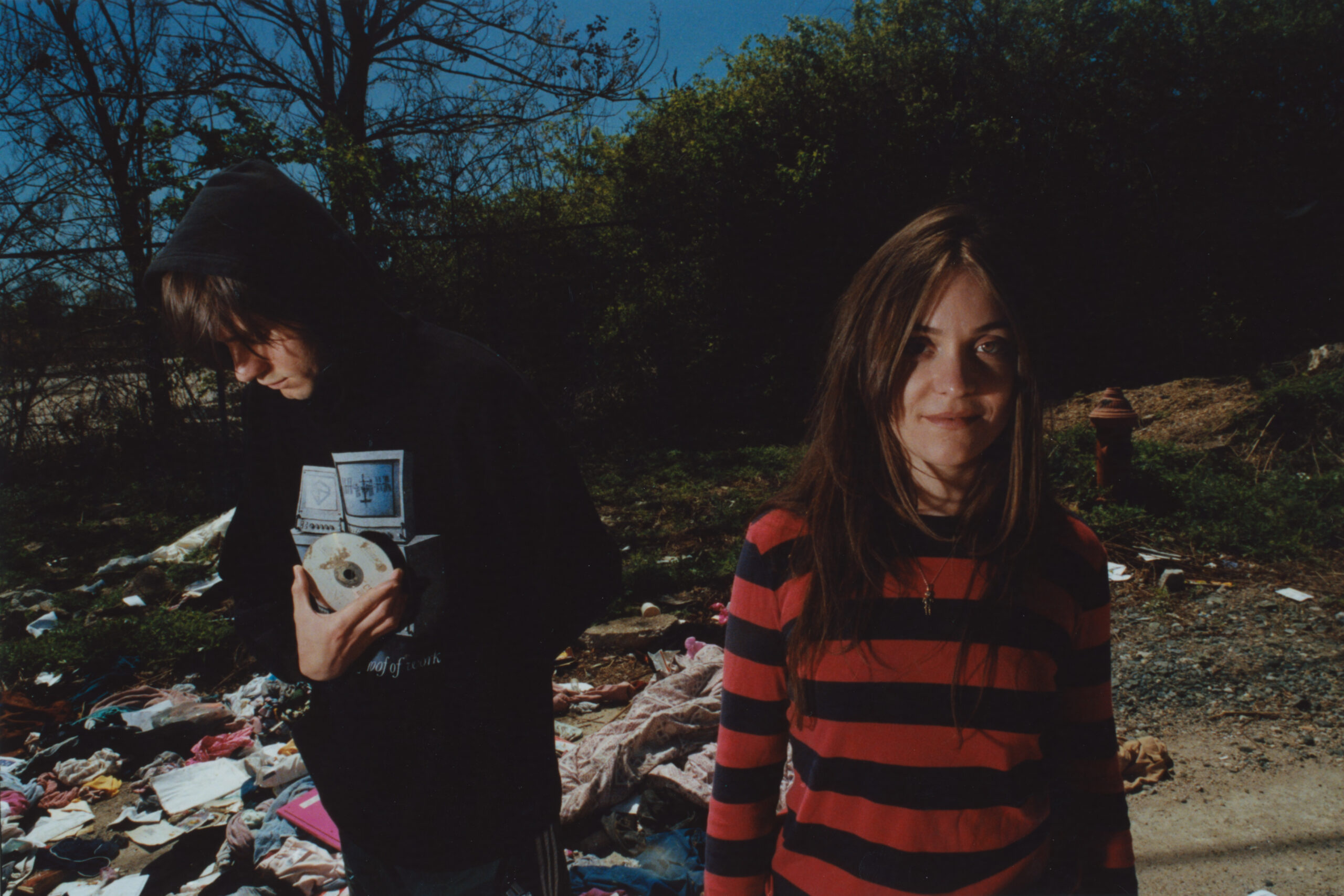 Two people are outdoors, standing on a littered ground with scattered debris and clothing. The person on the left is wearing a hoodie, holding an object close to their chest, while the person on the right is wearing a striped sweater and looking at the camera. Trees and a hydrant are visible in the background.