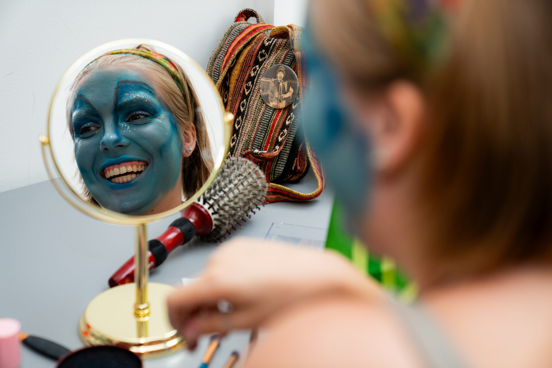 A photo taken over the shoulder of a person, visible in a mirror in the center of the screen. The person wears dramatic, blue makeup over their entire face.