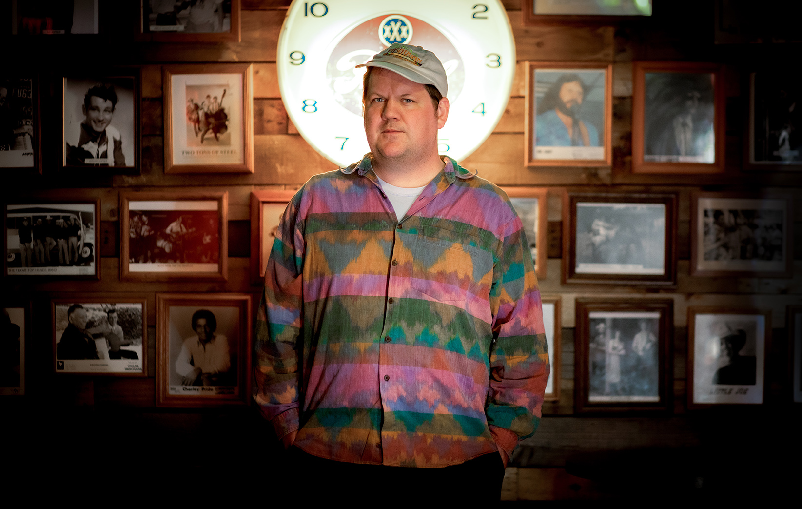 Jake Xerxes Fussell stands in a room with walls decorated with framed photos. A round white clock is behind him. He is wearing a colorful, patterned jacket and a cap.