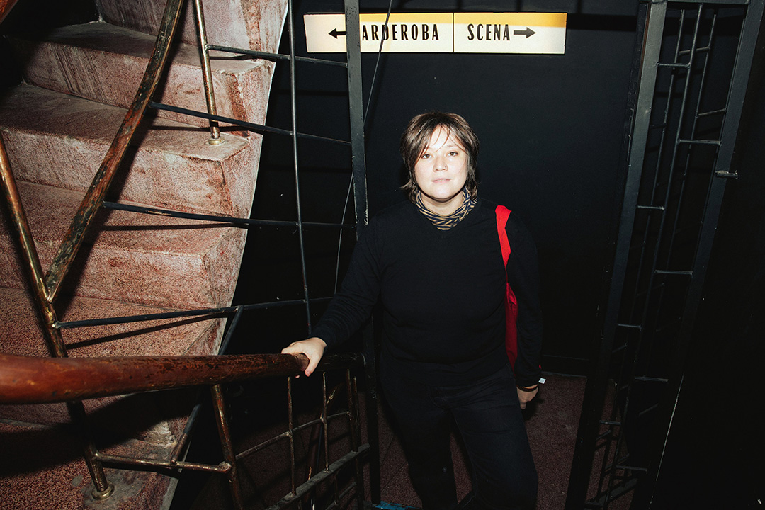 A person is standing on a dimly lit staircase, holding a railing. They are wearing a black outfit and have a red strap across one shoulder.