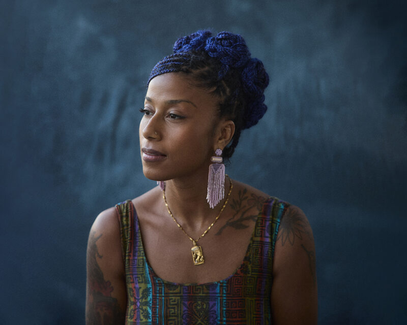 A person with braided blue hair is wearing a colorful patterned top, long tassel earrings, and a necklace. They have a thoughtful expression and tattoos on their arms. The background is a textured dark teal color.