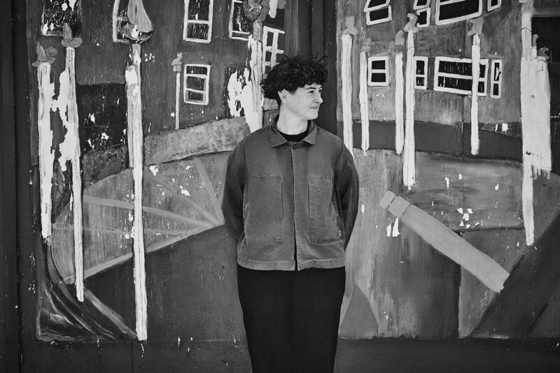 This black and white image shows a person standing in front of a mural with abstract shapes and distorted buildings. The mural features elongated white figures and a stair-like pattern. The person is looking to the side, with hands behind their back, wearing a jacket and dark pants.