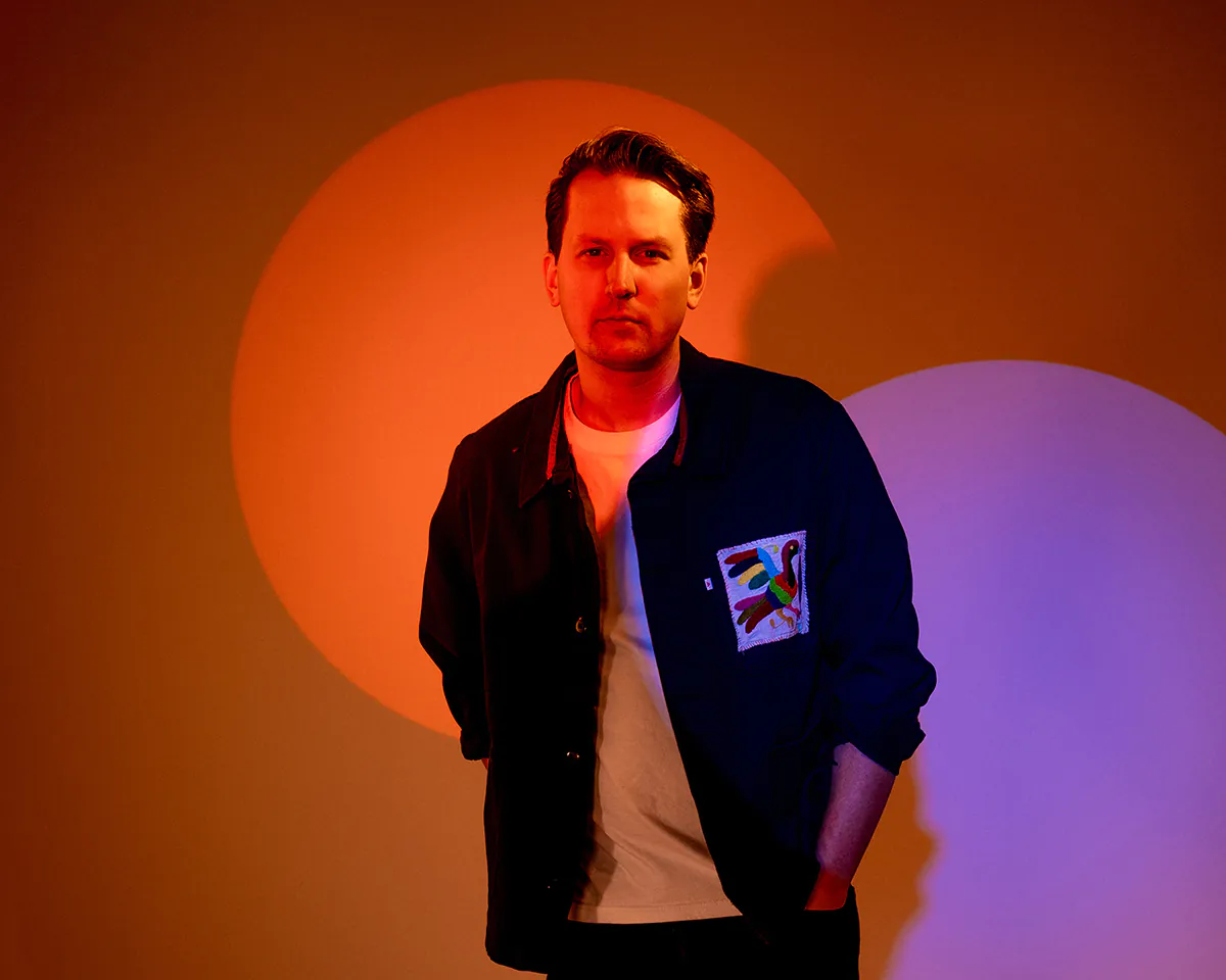 A person stands in a dimly lit room with colorful, overlapping circle lighting on the wall, wearing a dark jacket over a light shirt.