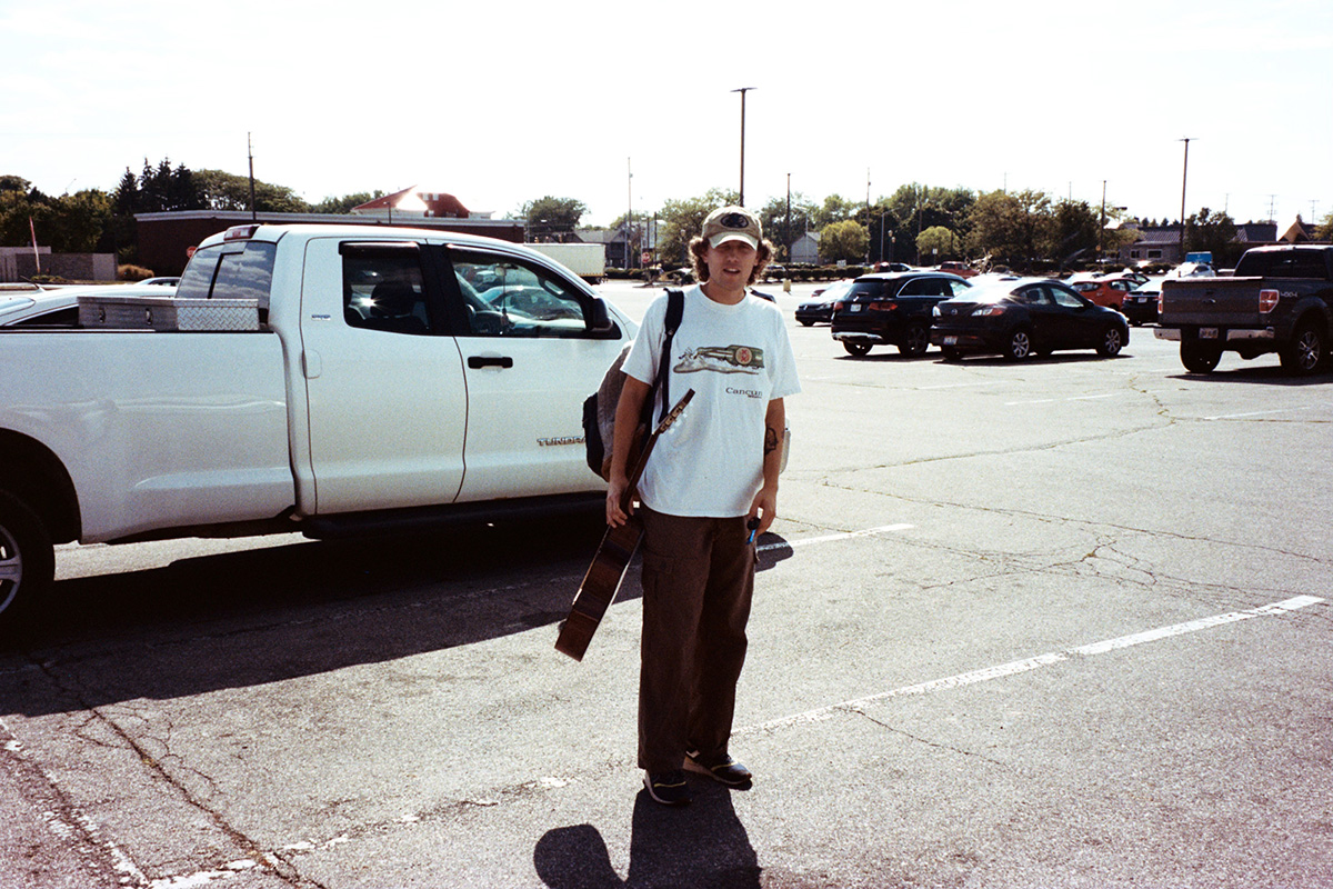 A person in a parking lot stands near a white pickup truck. The person is holding a guitar case and wearing casual clothes, including a cap. Several other vehicles are parked in the background under a clear sky.